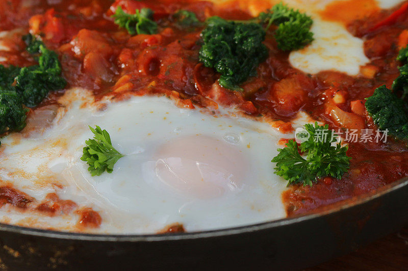 这是地中海健康早餐shakshouka / shakshuka / chakchouka的特写图片，用番茄、辣椒、洋葱、橄榄油和大蒜的酱汁煮熟的鸡蛋，上面撒上菠菜和欧芹